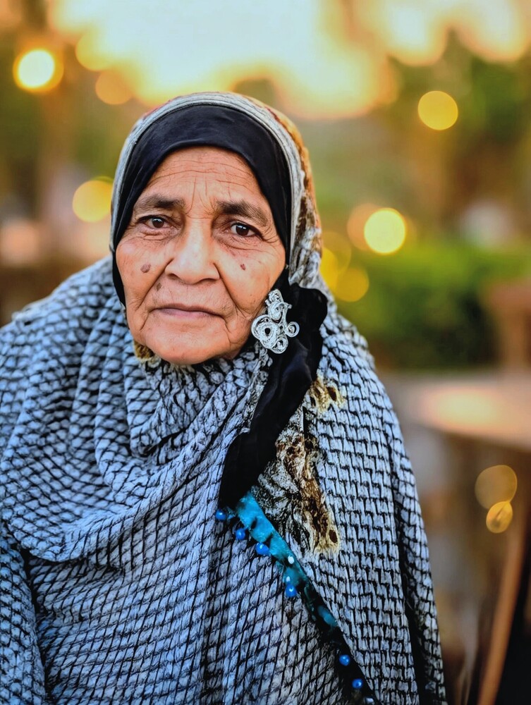 Elderly woman wearing light blue headscarf and embroidered black garment, warm golden hour lighting