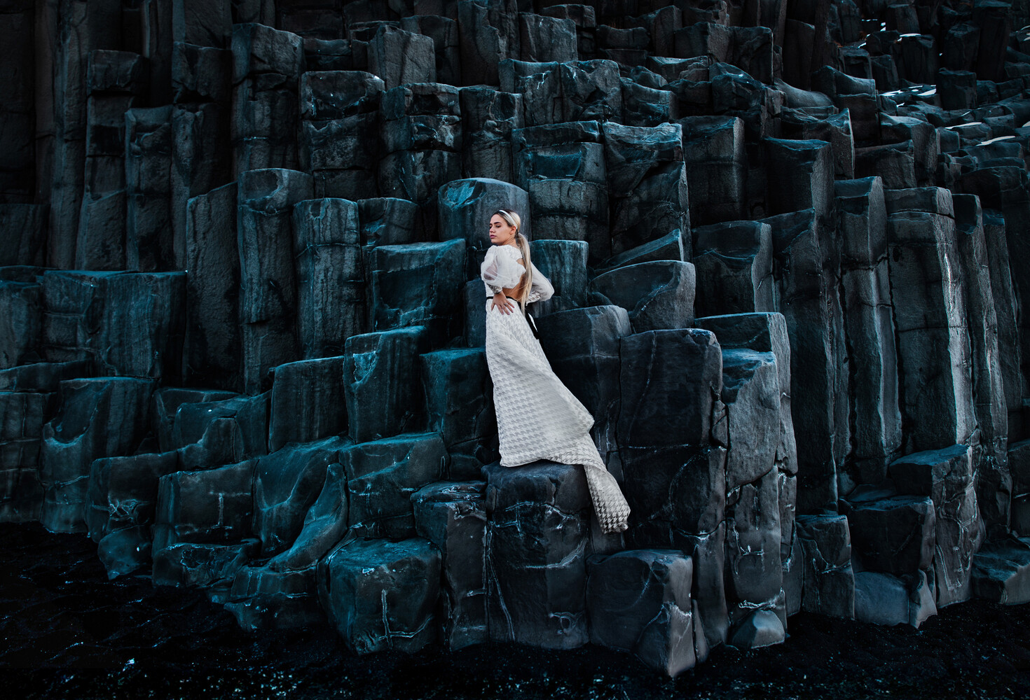 Woman in white draped clothing poses against dramatic dark basalt columnar rock formations
