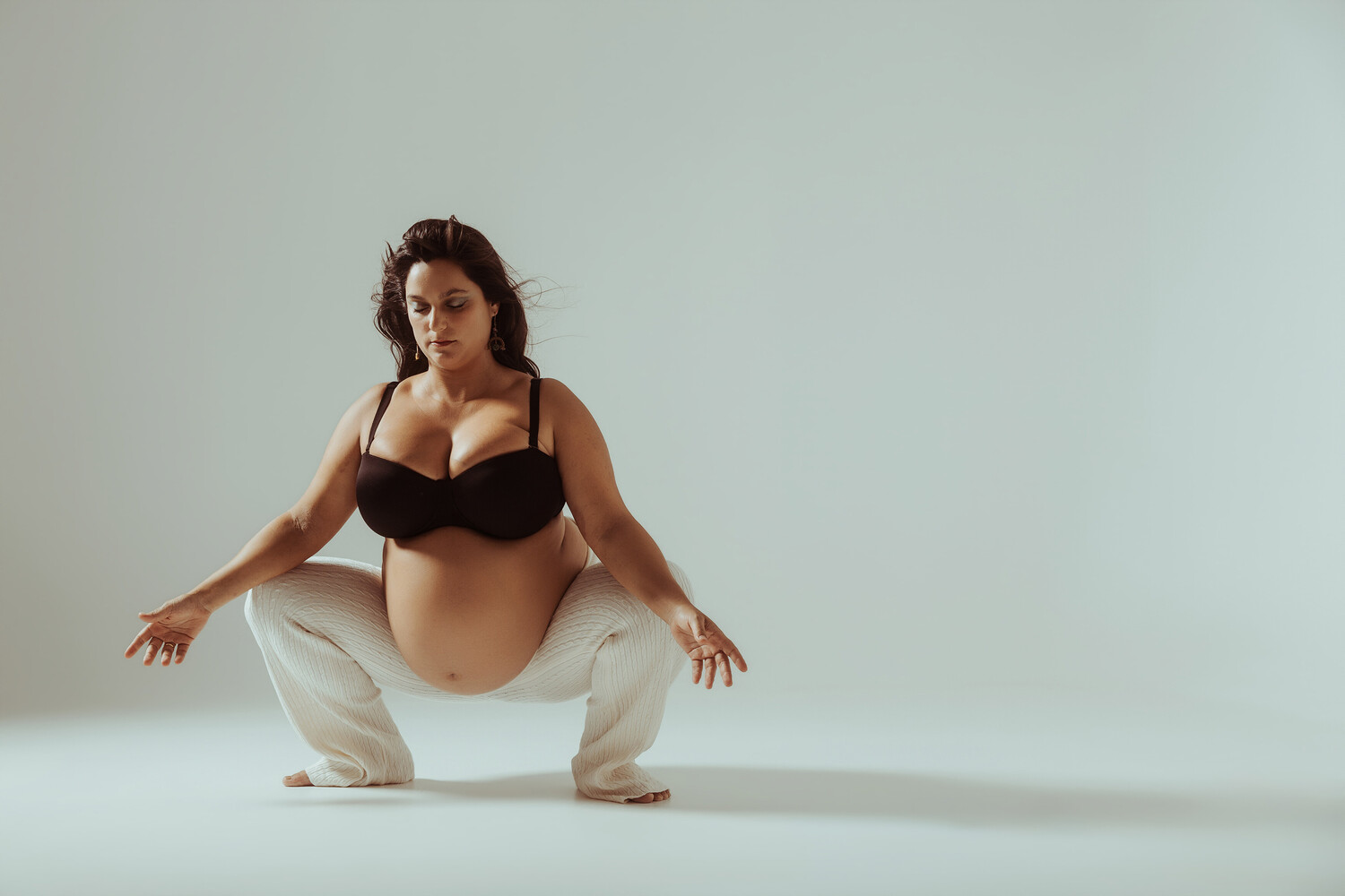 Woman in black bikini top and cream pants in squatting pose against neutral background