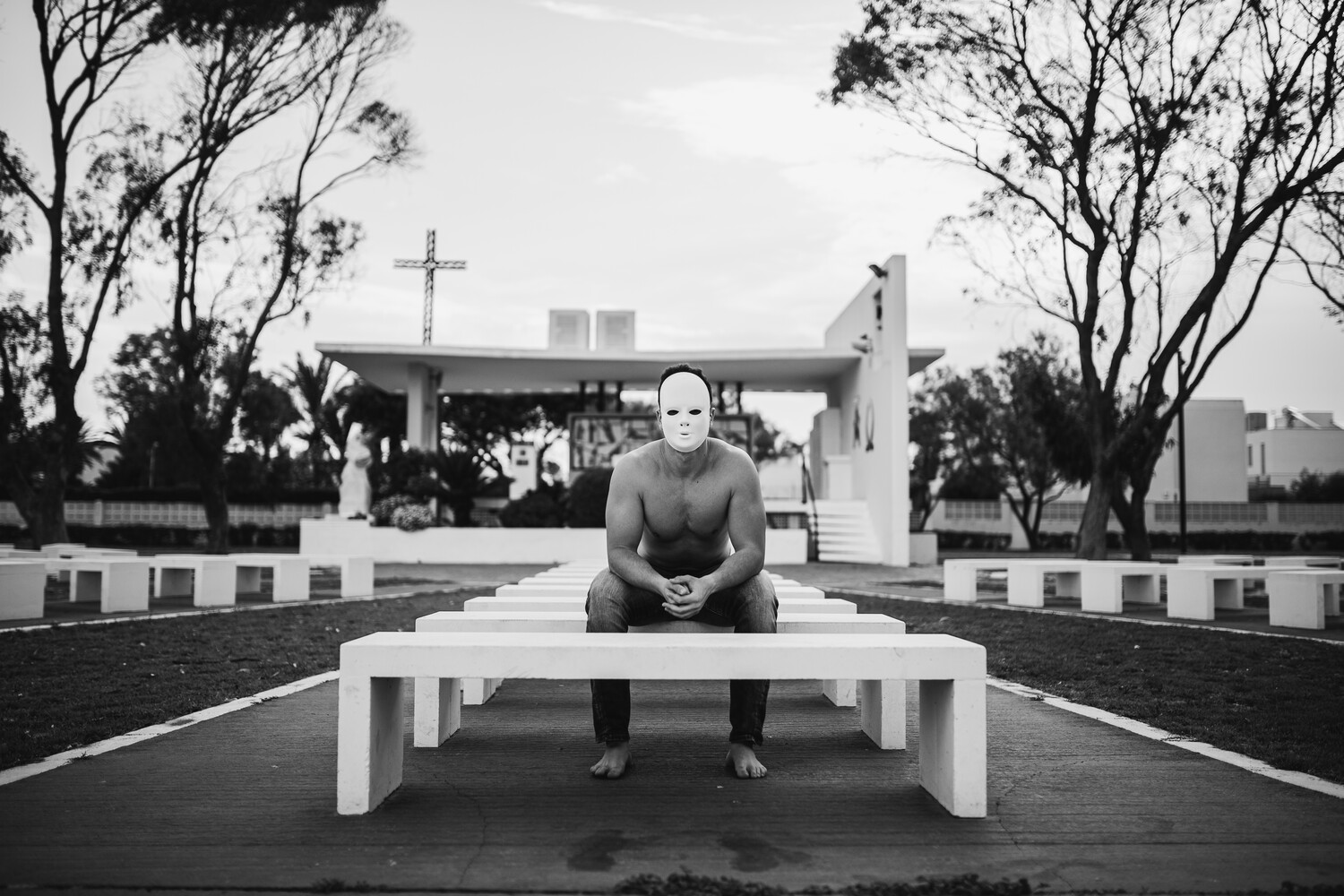 Shirtless man in mask sitting on white bench in symmetrical architectural courtyard