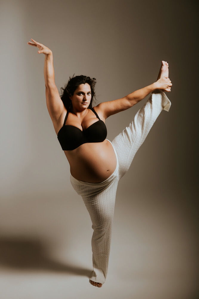 Pregnant woman in athletic wear performing a high leg kick pose against neutral background