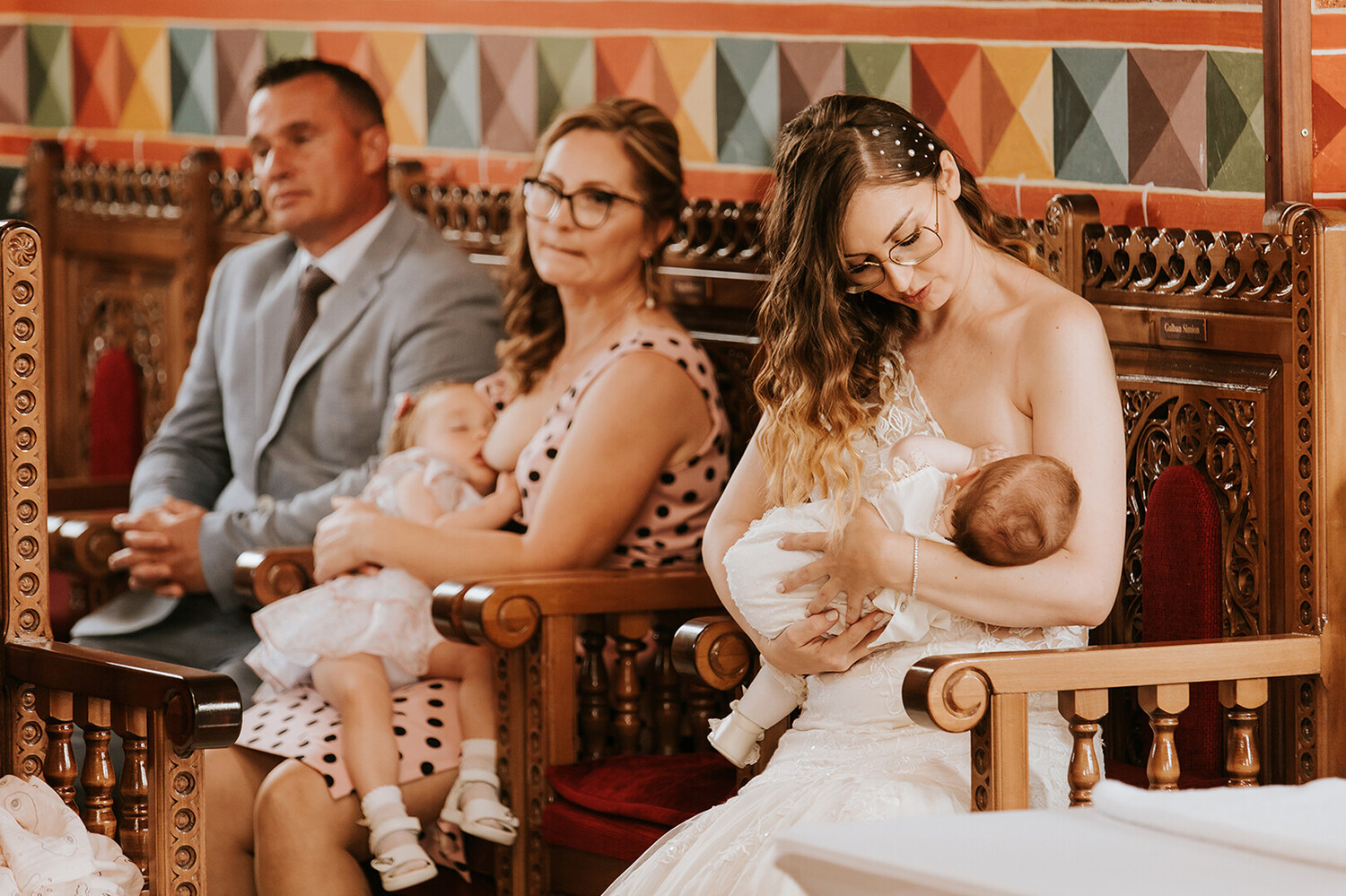 Woman in white wedding dress breastfeeding infant while seated with family members on ornate wooden furniture