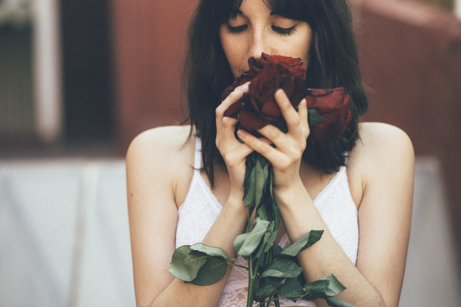 Woman with dark bob-cut hair, eyes closed, bringing a dark red rose to her face while holding it with both hands.