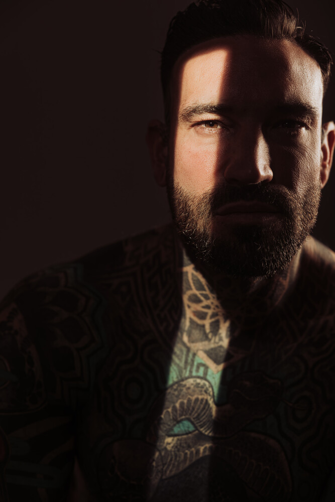 Bearded man with tattoos on his neck and chest, lit by harsh side light against a dark background.