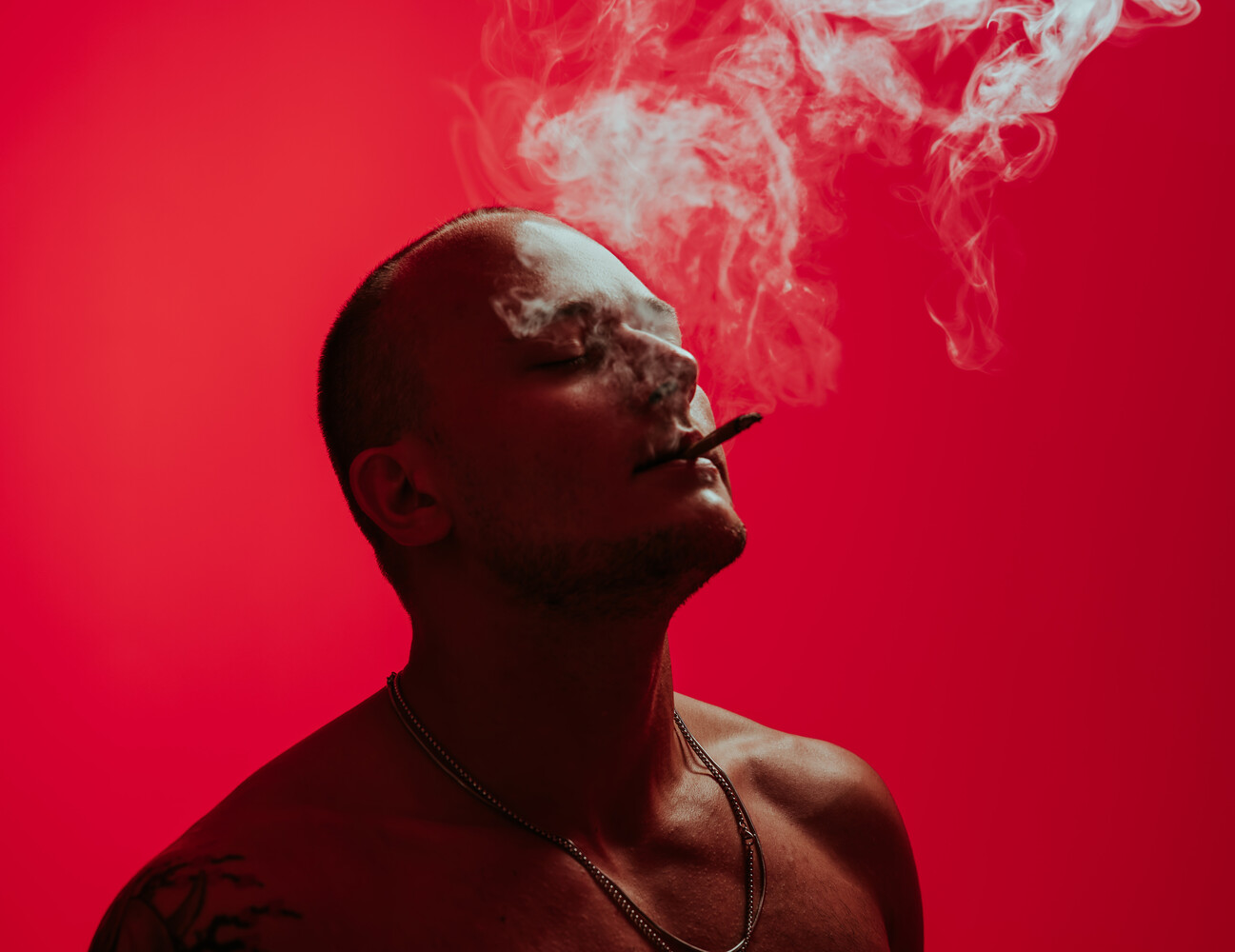 Shirtless man with a shaved head exhaling smoke from a cigar against a saturated red backdrop