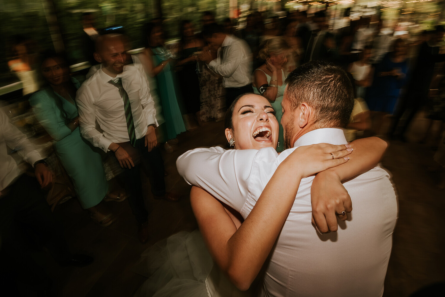 Bride in a white dress laughing with her eyes closed as she hugs a man in a white shirt, with guests blurred in the background.