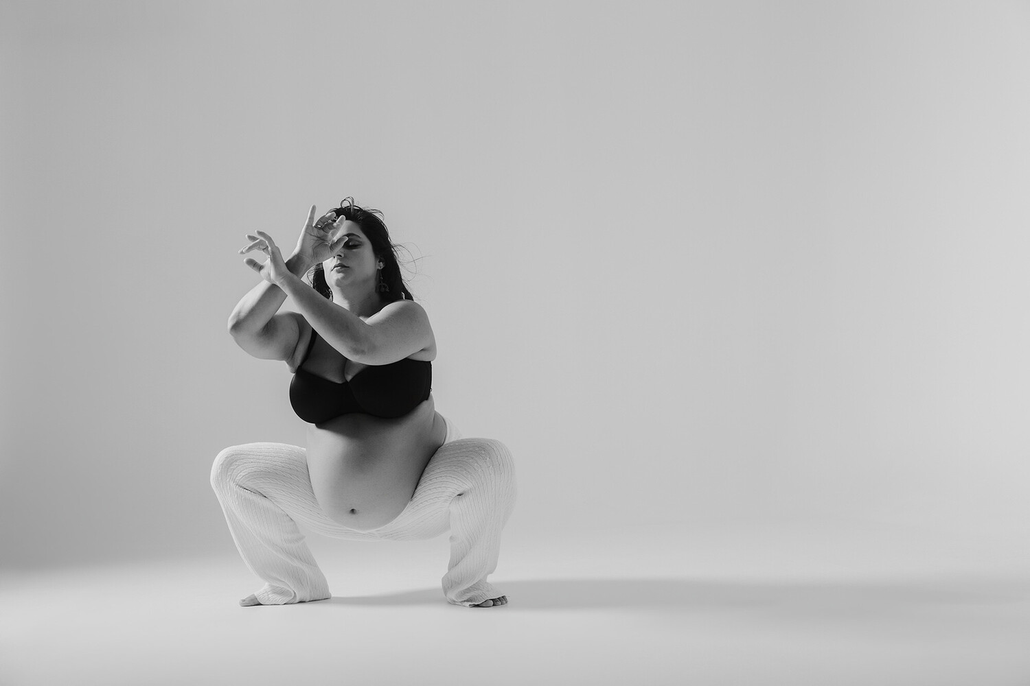 Black and white studio portrait of a pregnant woman in a crouched squat, arms raised and hands gesturing, wearing a black sports bra and white pants against a seamless backdrop.