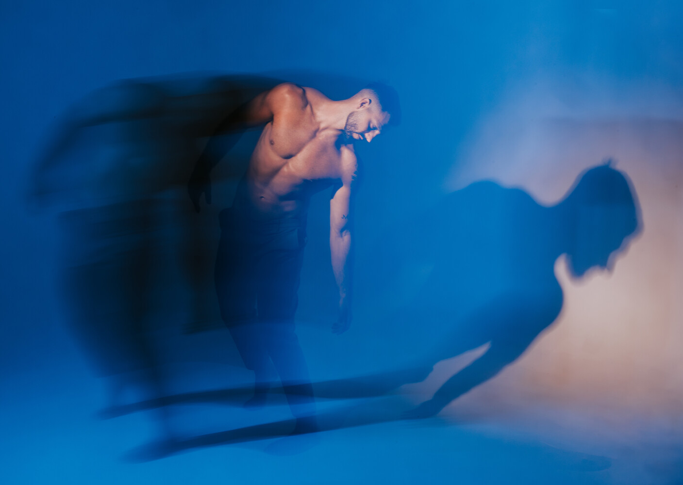 Shirtless dancer in motion blur leaning forward against a deep blue backdrop, with a long shadow cast on the wall.