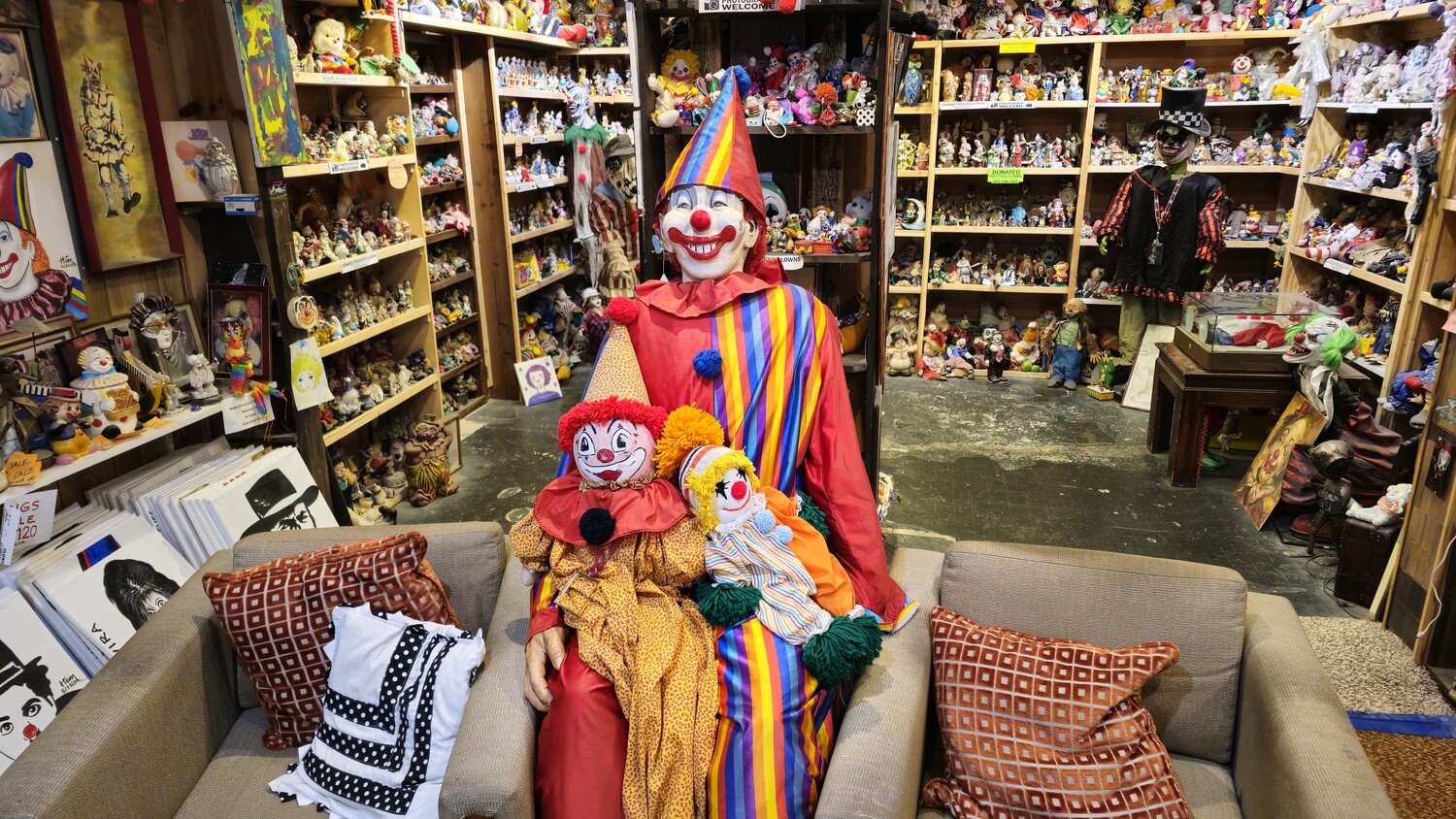 Life-sized clown figure in red costume with striped collar holding smaller clown dolls on couch in collectibles shop
