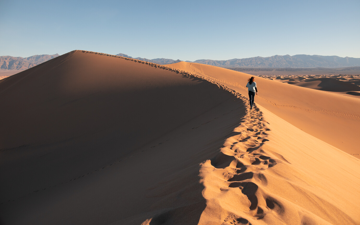 Hiker ascending a massive sand dune with footprints trailing behind in golden afternoon light