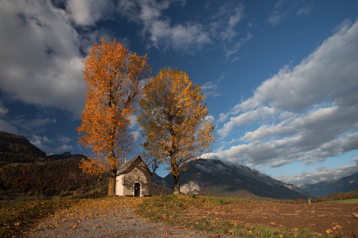 Small stone chapel with a steep roof beside two tall autumn trees with orange-gold leaves, set against mountains under a blue sky with clouds.