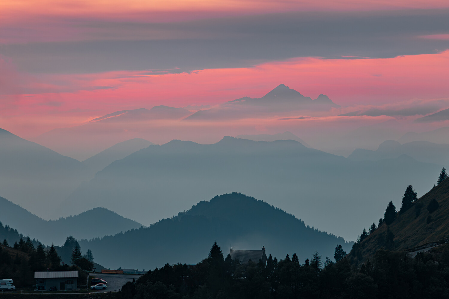 Layered misty mountain ridges under a pink twilight sky, with a distant snowy peak and dark conifers in the foreground.