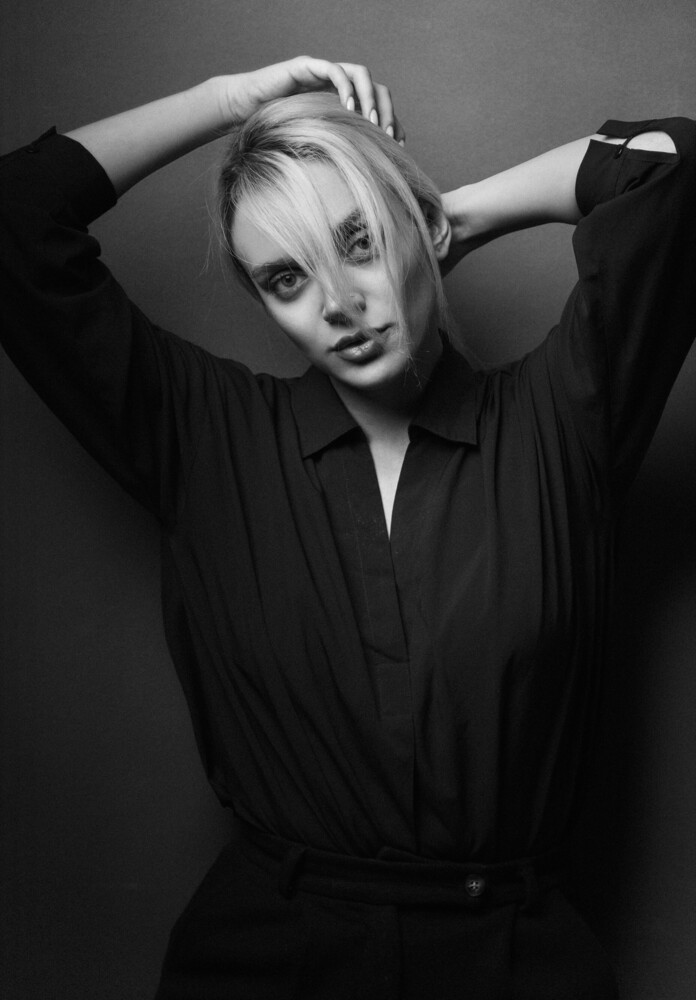 Woman with blonde hair in black robe, hands raised to head, posed against neutral background