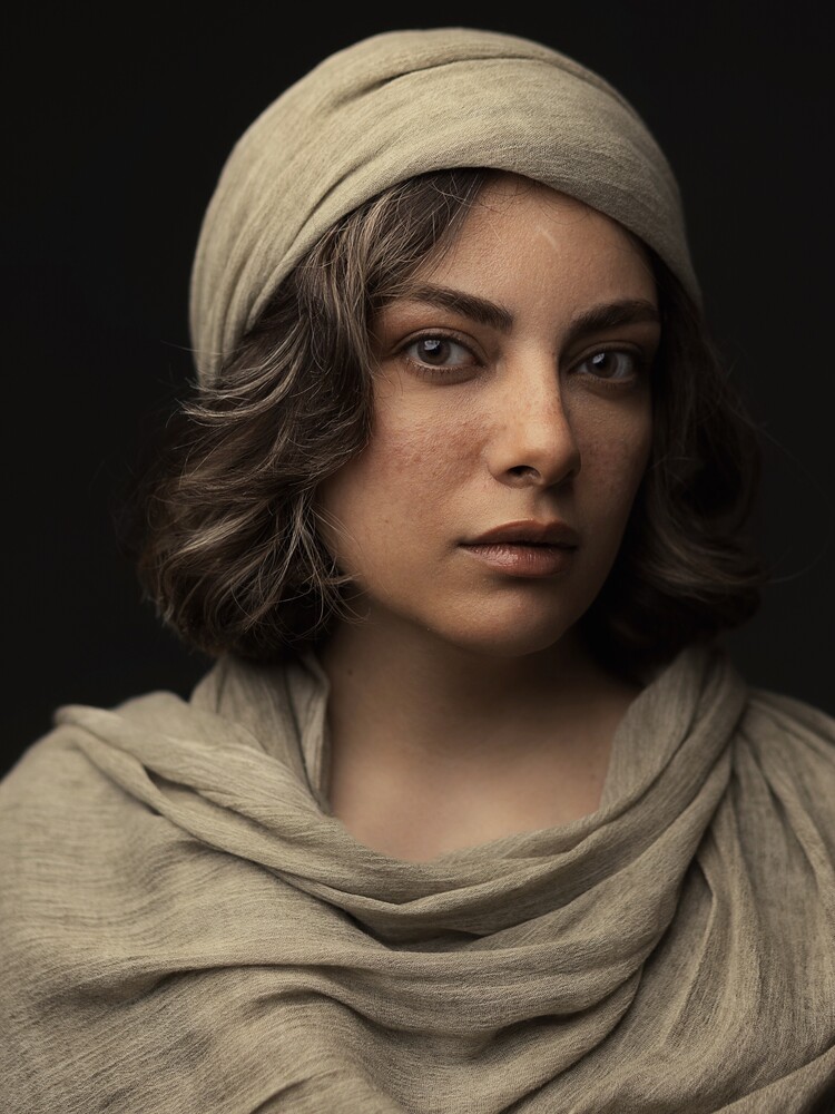 Close-up portrait of a woman with freckles, wearing a beige linen headwrap and draped scarf against a dark background, gazing directly at the camera.