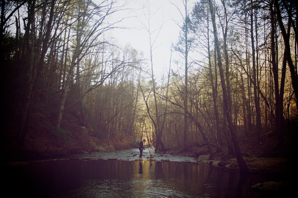 Person standing in shallow stream surrounded by bare deciduous trees in misty forest