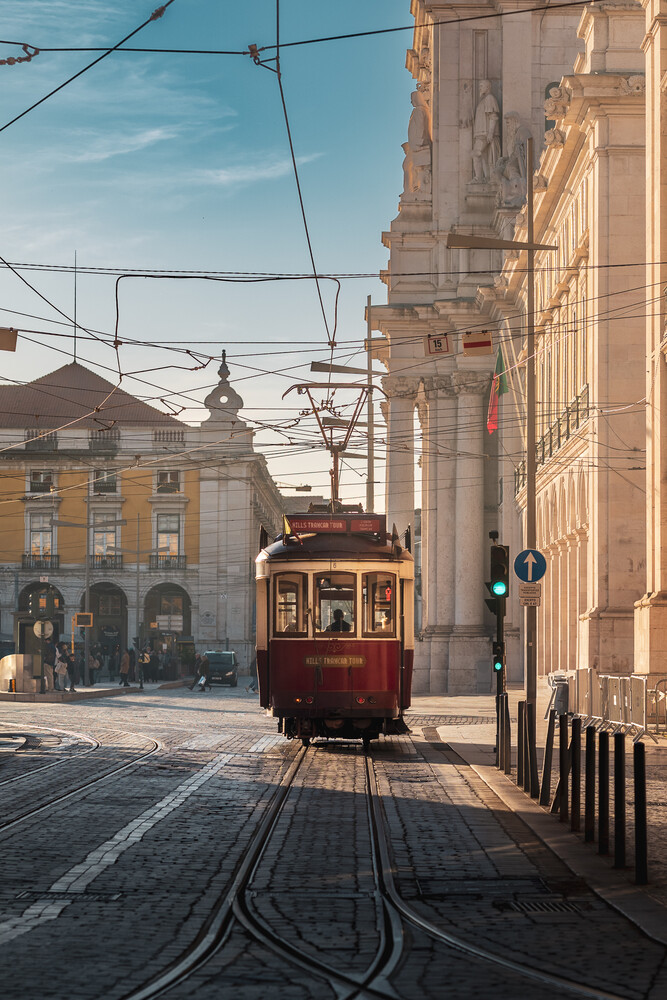Red vintage tram rolling toward the camera down a sunlit street with tracks and historic buildings.