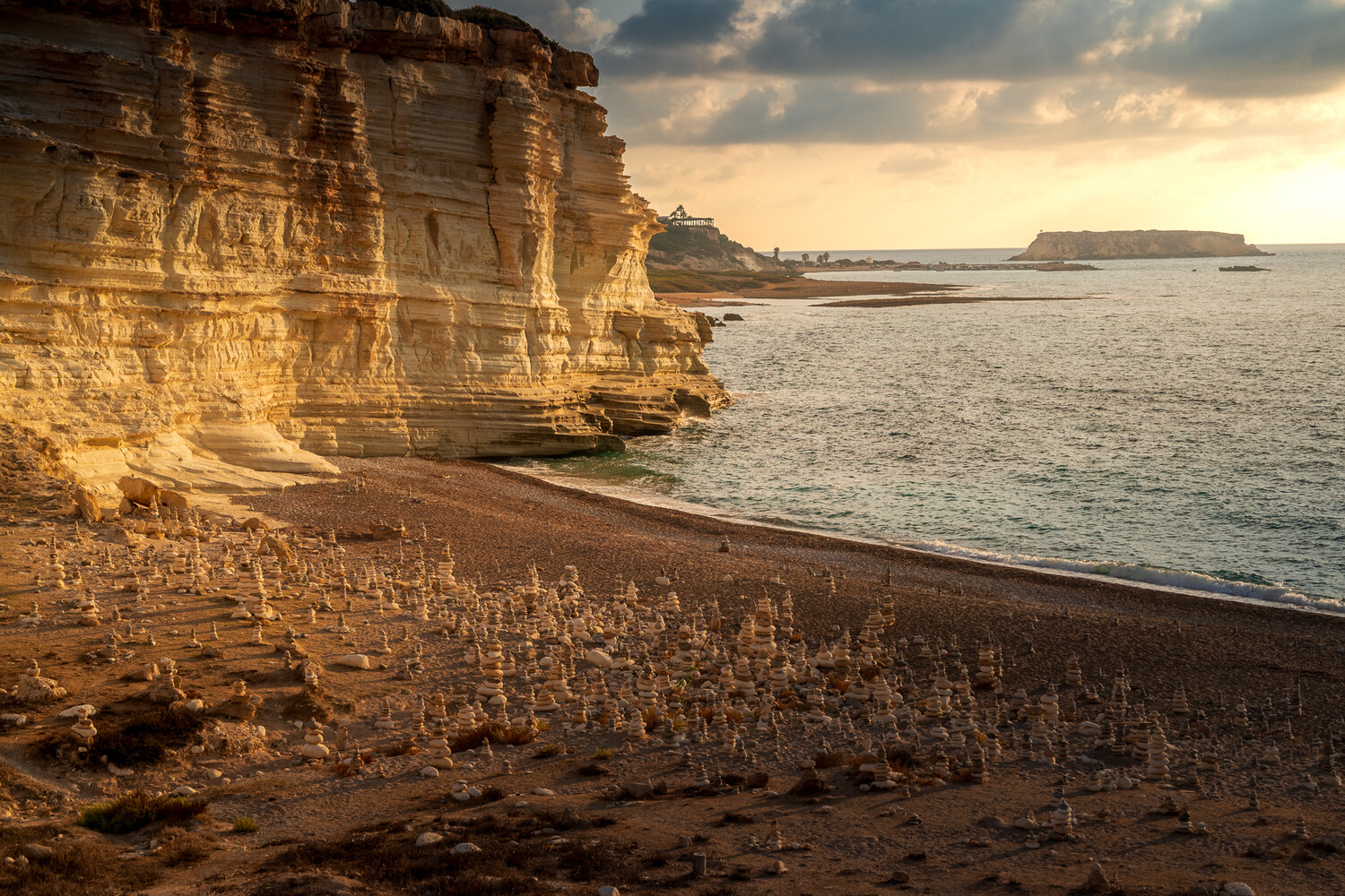 Layered golden sandstone cliff lit by warm sunset light beside a calm sea, with a rocky headland in the distance.