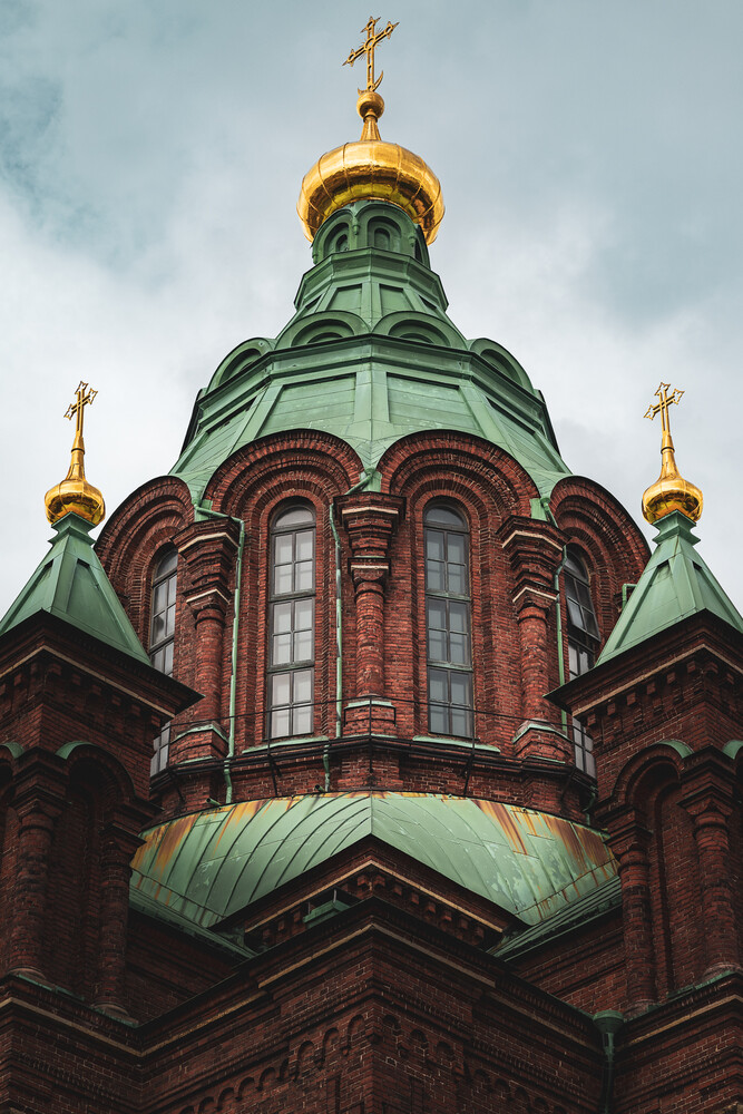 Low-angle view of a red brick church with green copper domes and gilded onion domes topped with gold crosses against a cloudy sky.