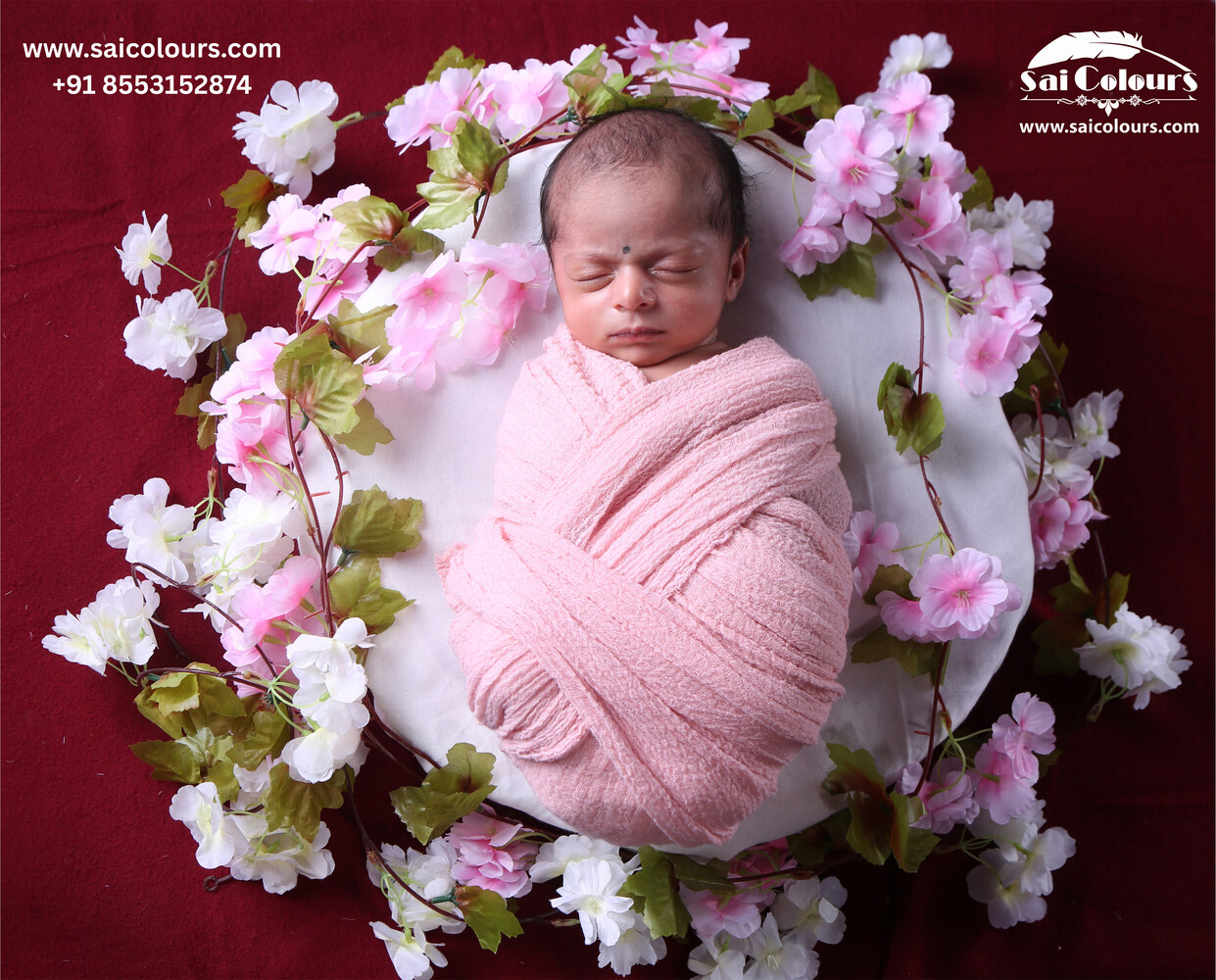 Newborn baby asleep wrapped in pink fabric on a white dish, framed by pink and white artificial blossoms on a red backdrop.