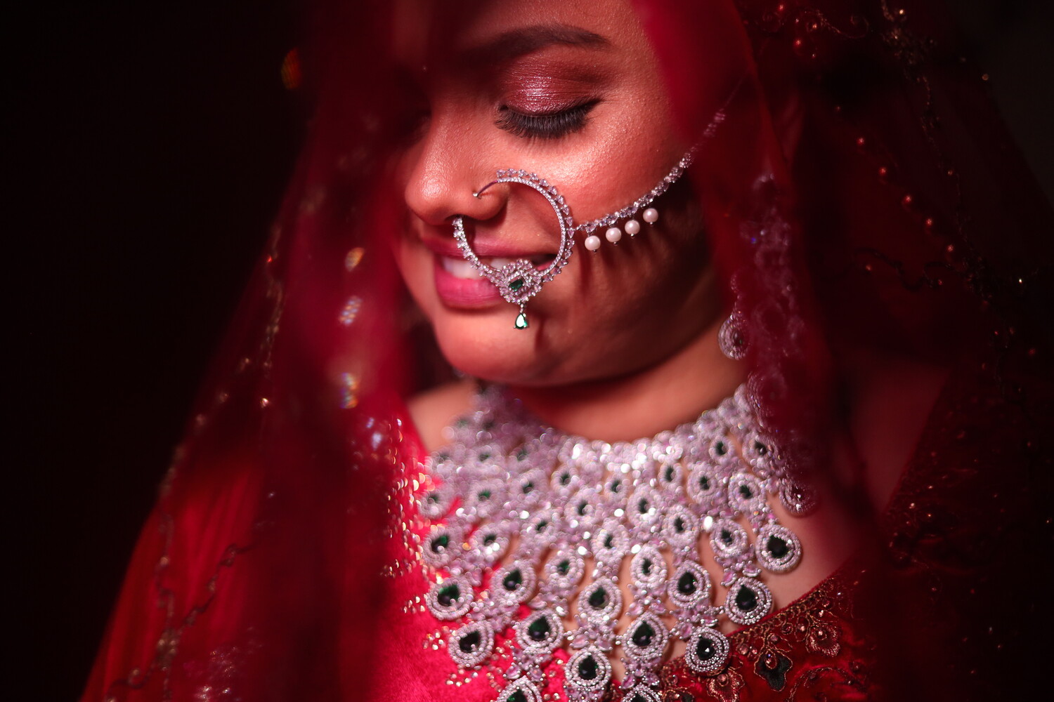 Woman with lowered eyes in a red veil wearing an ornate nose ring and embellished bridal necklace on a dark background