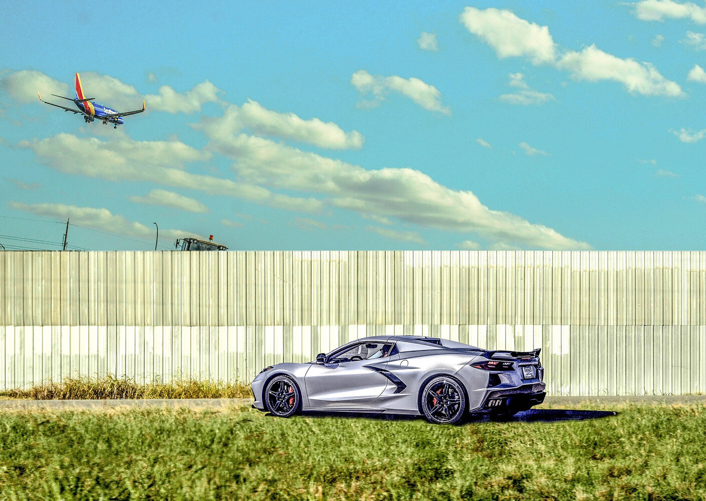 Silver mid-engine Corvette parked on grass by a tall fence, with a jet in Southwest colors descending overhead