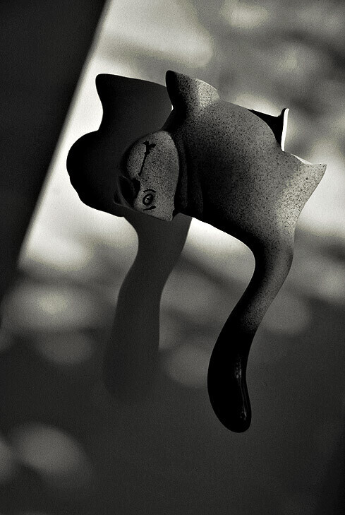 Black-and-white photo of a fish-shaped figurine with a curved tail, casting a dark shadow against a bright, dappled background