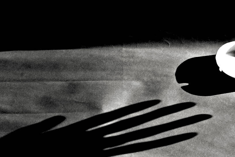 Black and white photo of a spread hand's shadow stretching across a pale surface toward a bright light source at the right edge