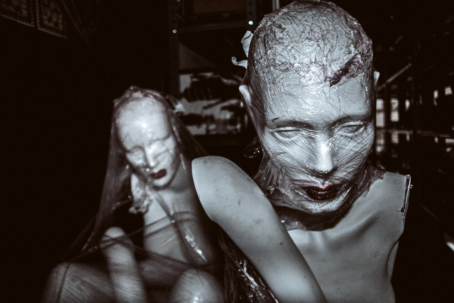 Black-and-white photo of two pale mannequin-like figures wrapped in clear plastic film against a dark background, the front figure's face in sharp focus.