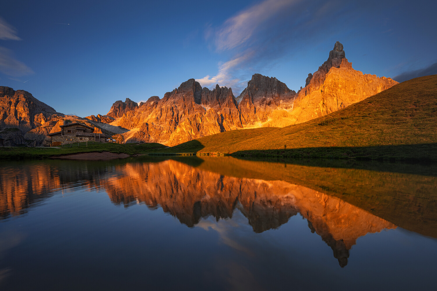 Sunlit jagged alpine peaks reflected in a calm mountain lake at golden hour, with a rustic stone hut on the left shore.