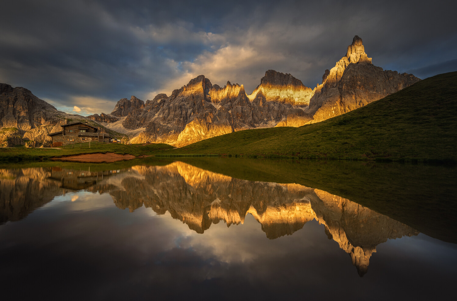 Golden-lit jagged mountain peaks reflected in a still alpine lake, with a wooden mountain hut on a grassy shore beneath stormy clouds.