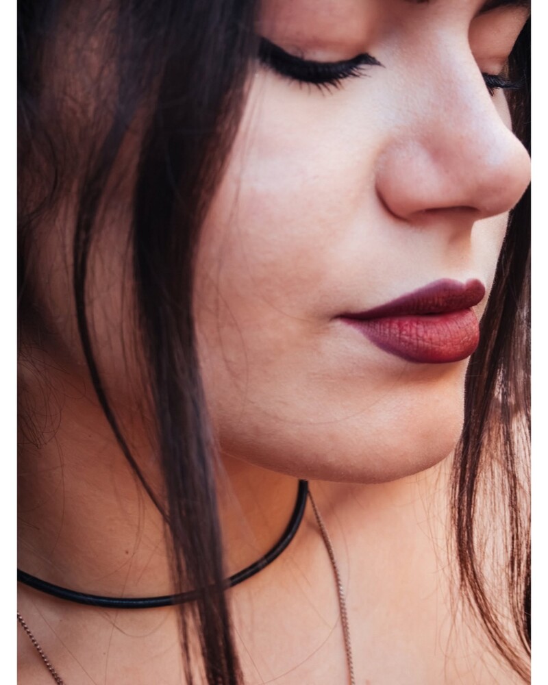 Close-up portrait of a woman with eyes lowered, wearing dark burgundy lipstick and a thin black choker necklace, dark hair falling across her shoulder.
