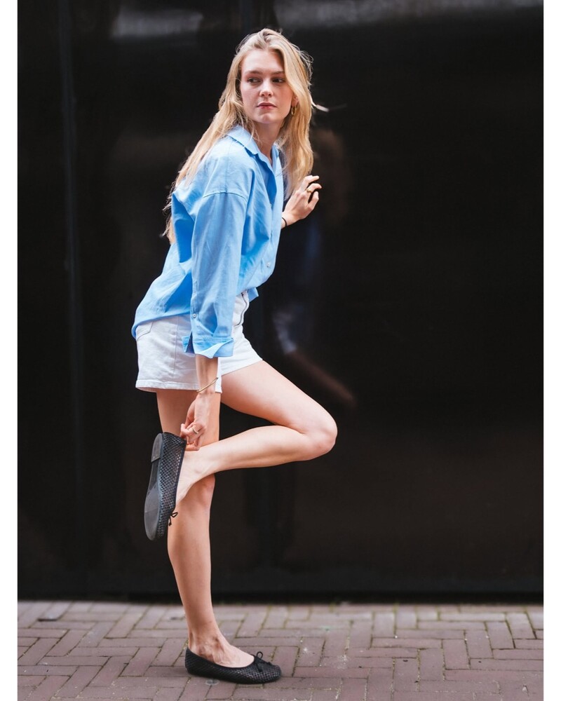 Woman with long blonde hair in a light blue button-up shirt and white shorts, balancing on one leg against a dark wall