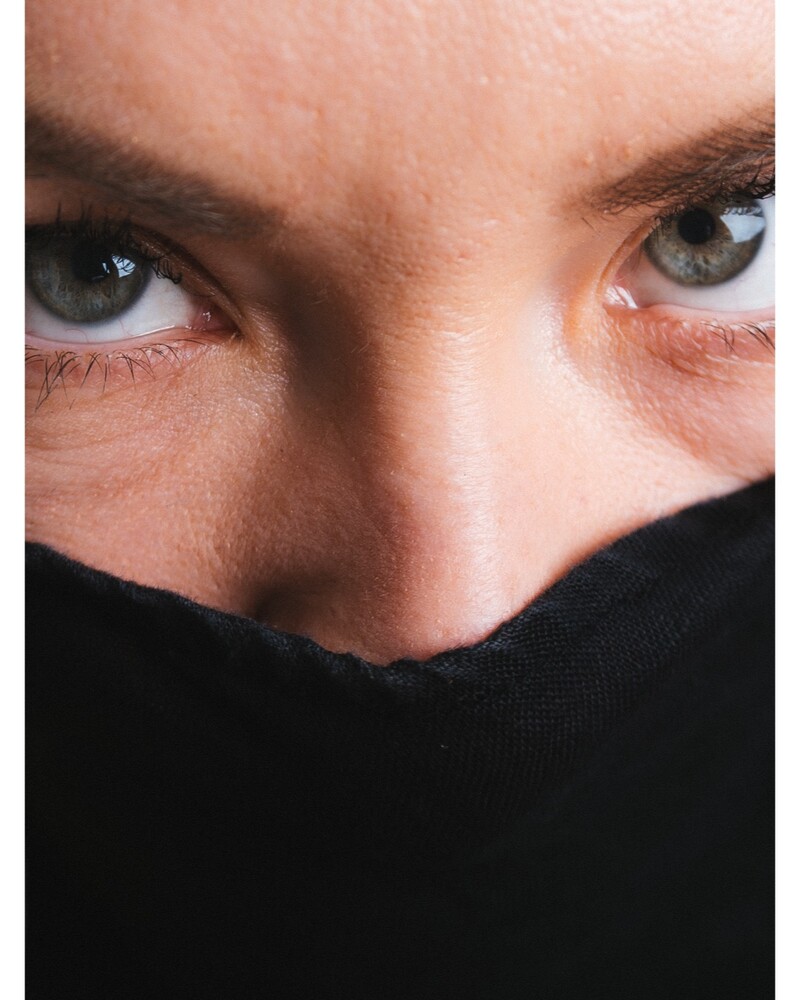 Close-up portrait of a woman's grey-green eyes above a black fabric covering her nose and mouth.