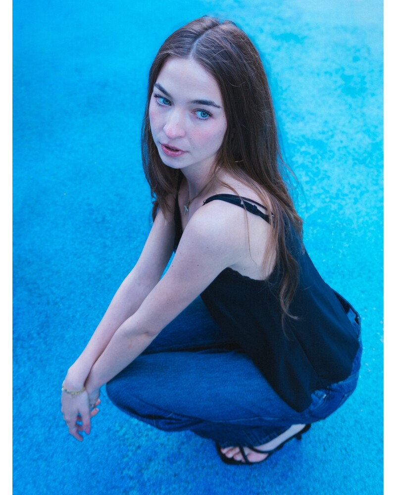 Woman with long brown hair in a black camisole and blue jeans crouching on a vivid blue textured surface, looking up at the camera.