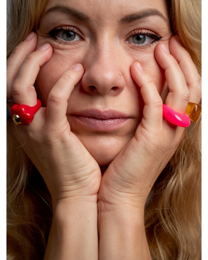 Close-up portrait of a blonde woman with blue eyes cupping her face in both hands, wearing bright red and pink rings.