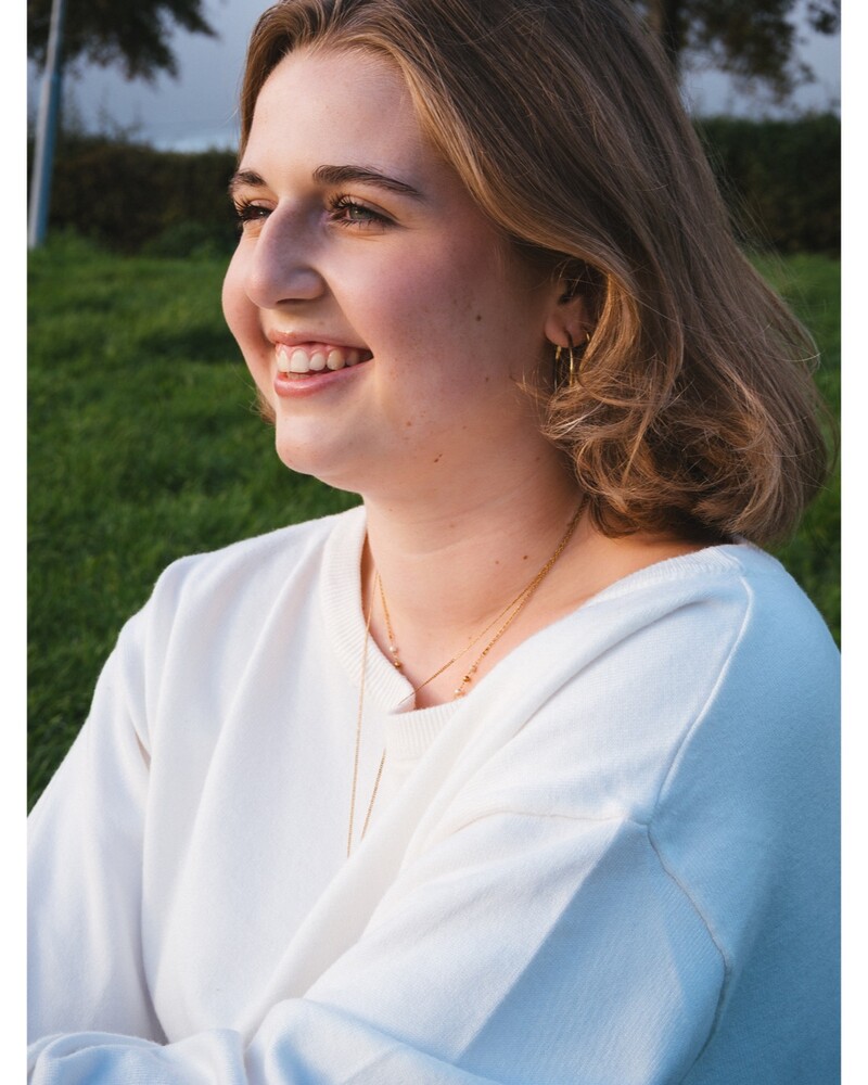 Smiling woman with shoulder-length wavy light brown hair, wearing a white sweater and gold necklace, looking off to the side against a blurred green lawn.
