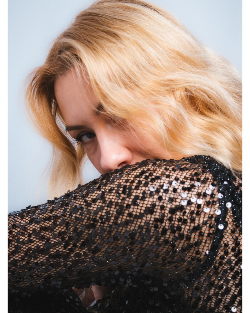 Woman with wavy strawberry-blonde hair leaning her face into the sleeve of a black sheer sequined top against a pale blue backdrop.