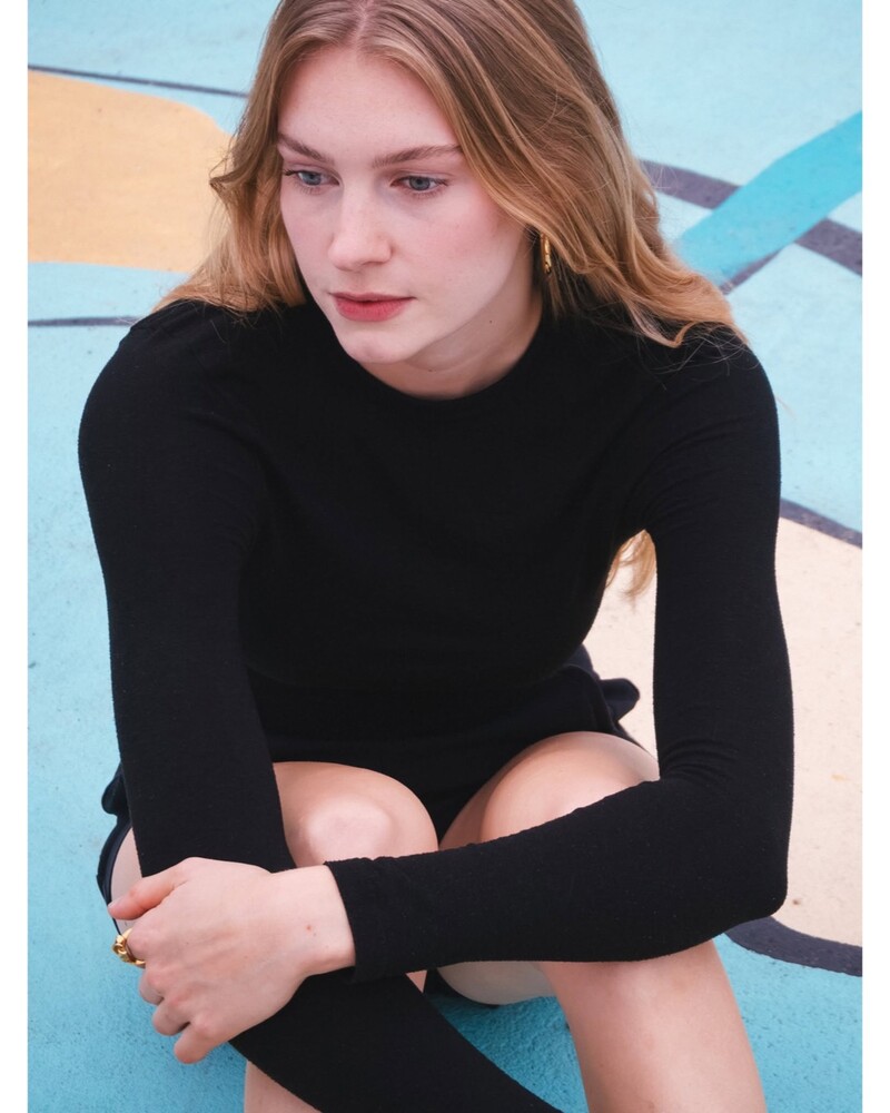 Woman with long blonde hair in a black long-sleeve top sitting with knees up against a blue and yellow painted wall.