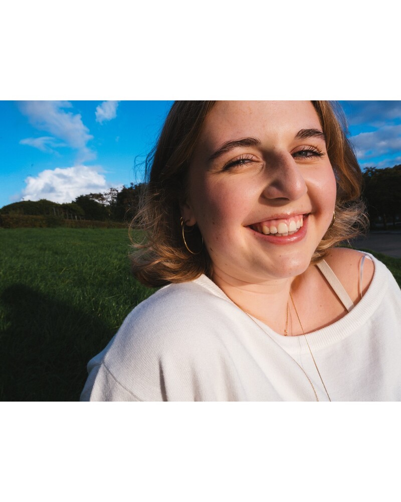 Smiling woman with shoulder-length hair and a hoop earring, lit by warm sunlight in a grassy field under a blue sky