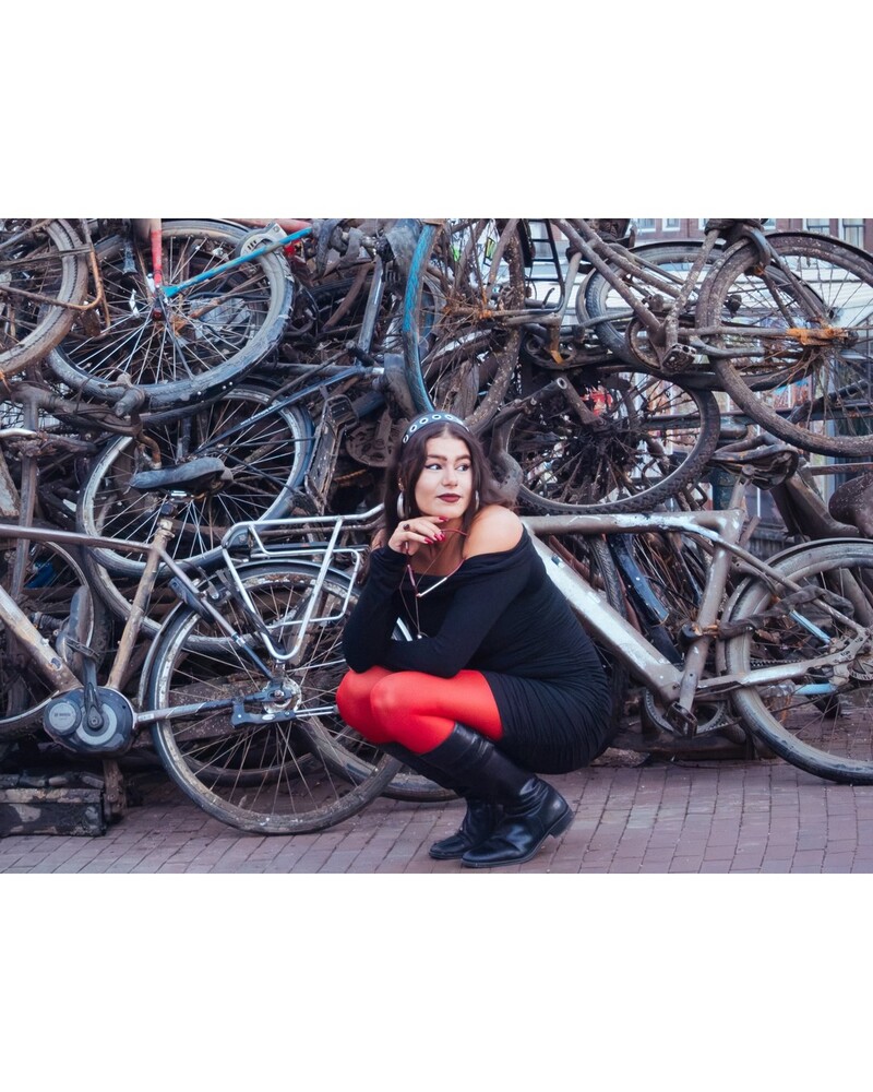 Woman in black outfit and red tights crouching in front of a tangled pile of old bicycles on a brick street