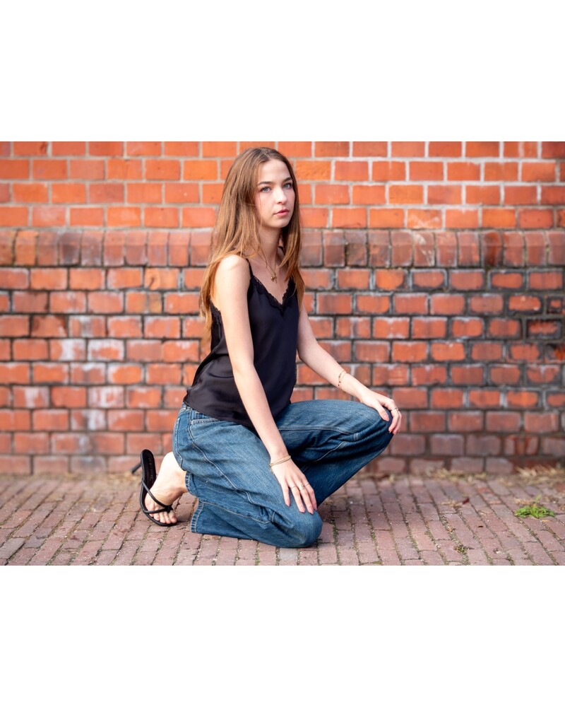 Woman with long brown hair in a black camisole and dark jeans crouching on a brick pavement in front of a red brick wall.