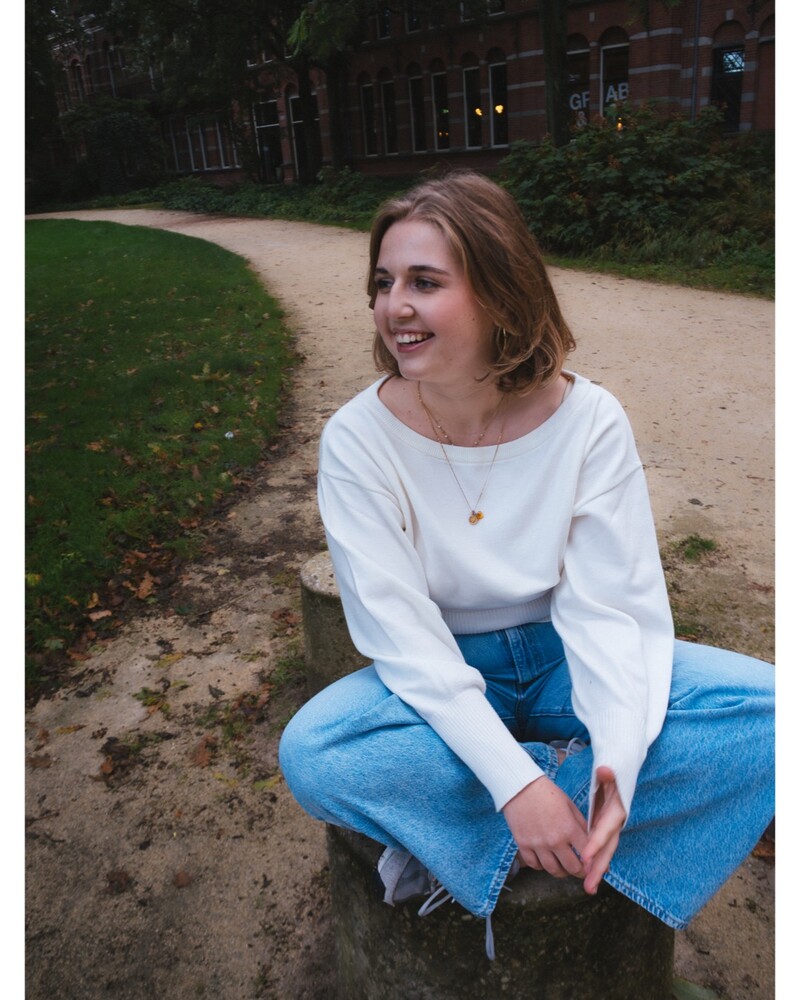 Smiling woman with shoulder-length hair in a white top and blue jeans, sitting on a stump beside a gravel path