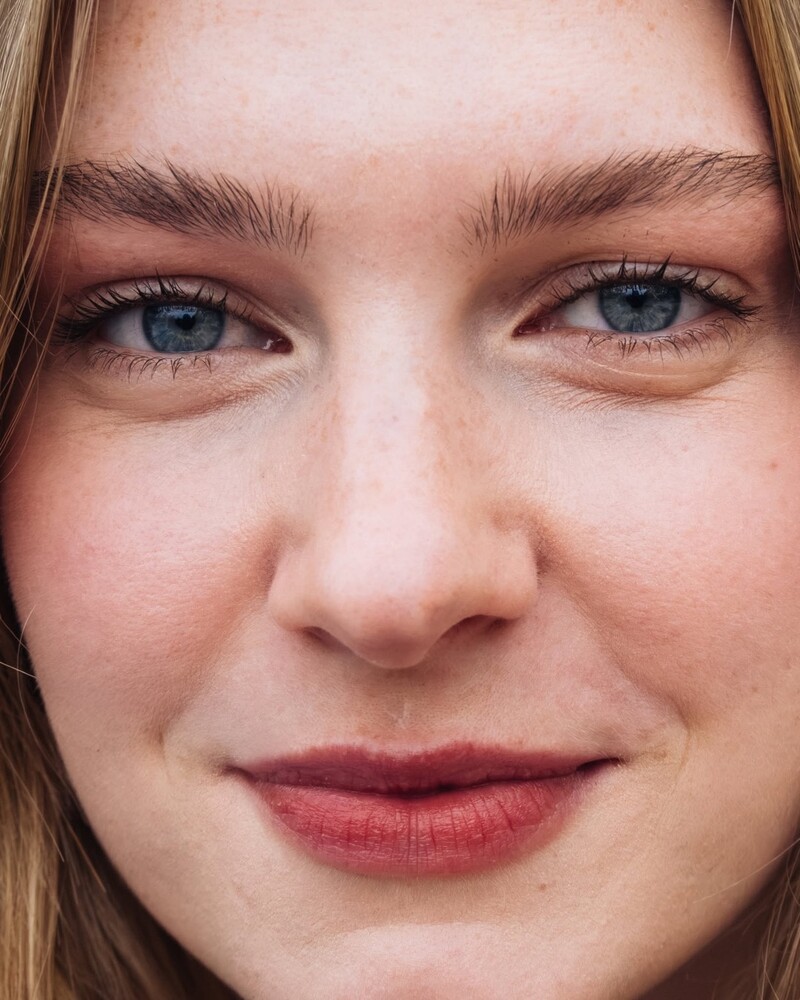 Close-up portrait of a young woman with light blue eyes, blonde brows, and freckled skin, lightly smiling with a rosy-red lip.