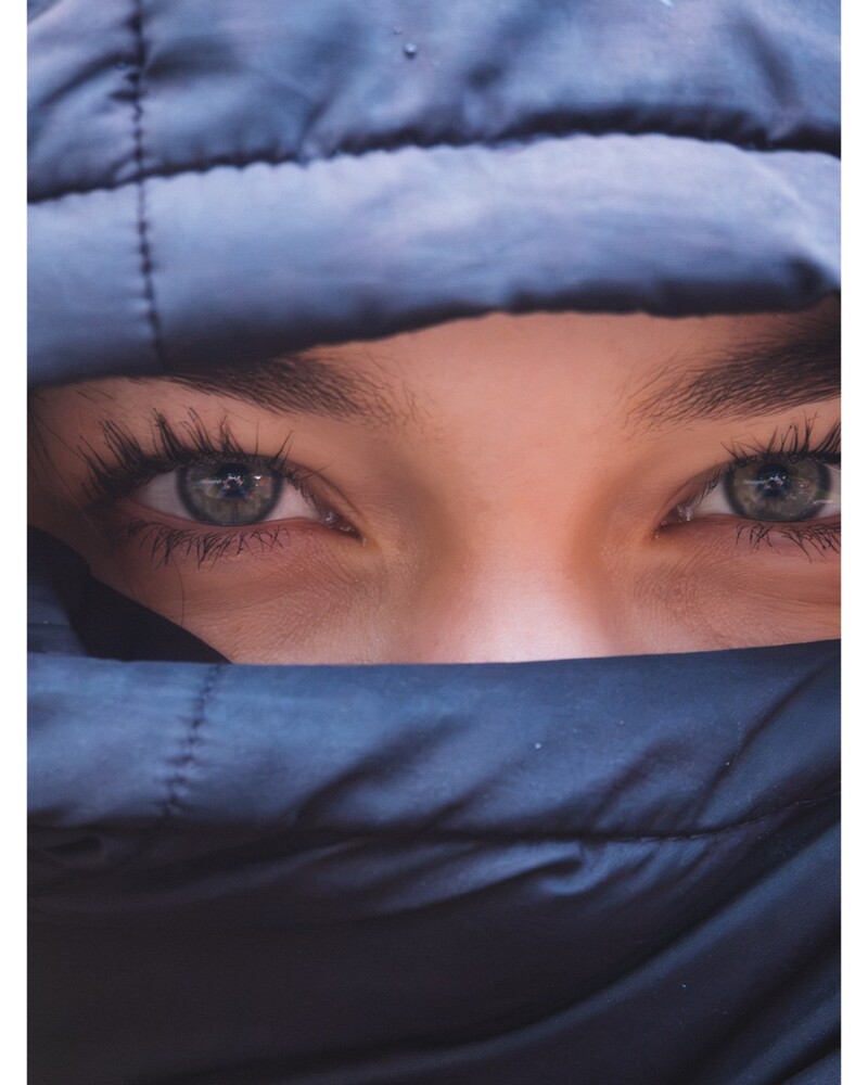 Close-up portrait of a person's light blue-gray eyes, framed by a dark blue quilted fabric wrapped over the lower face.