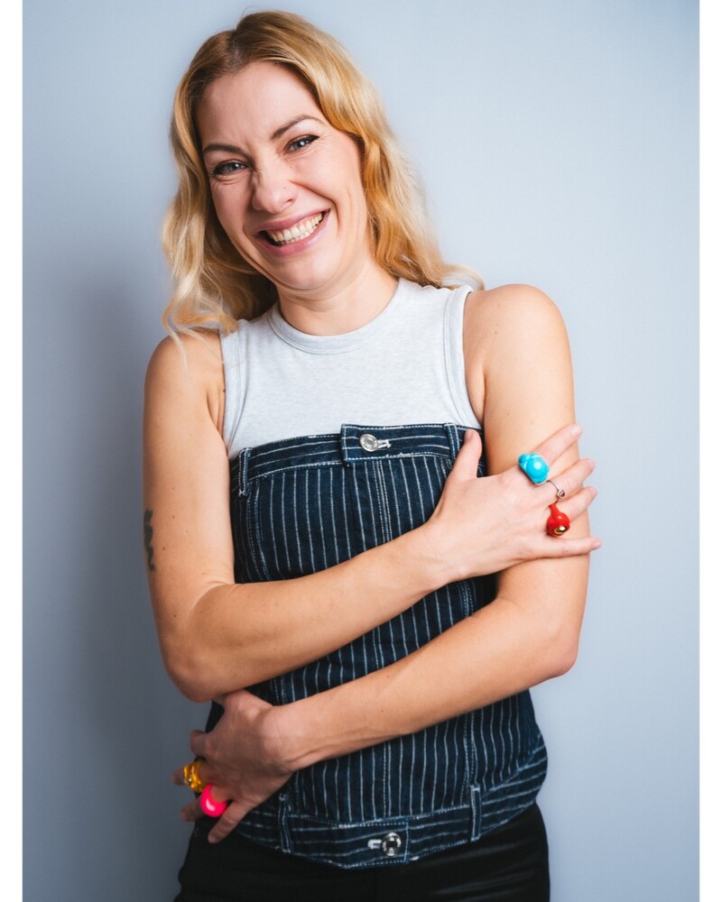 Smiling blonde woman in pinstripe overalls and a grey tank top, arms crossed, against a plain grey backdrop