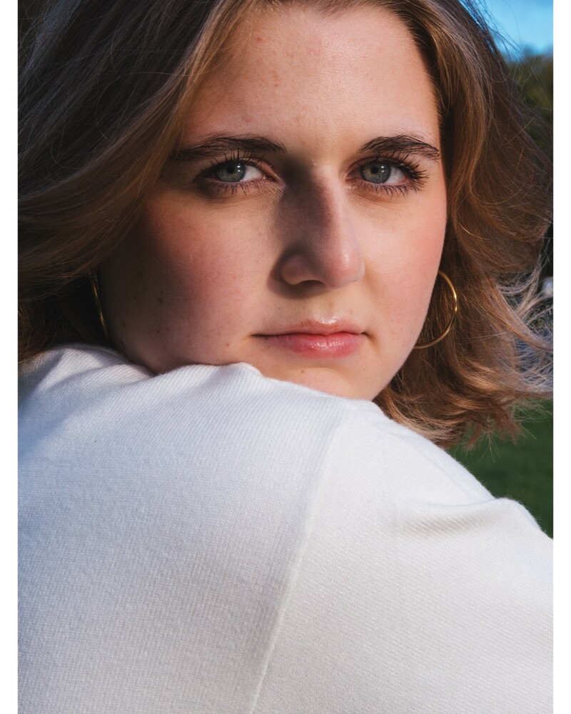 Young woman with wavy light-brown hair and gold hoop earrings, resting her chin on a white sweater shoulder and gazing toward the camera.