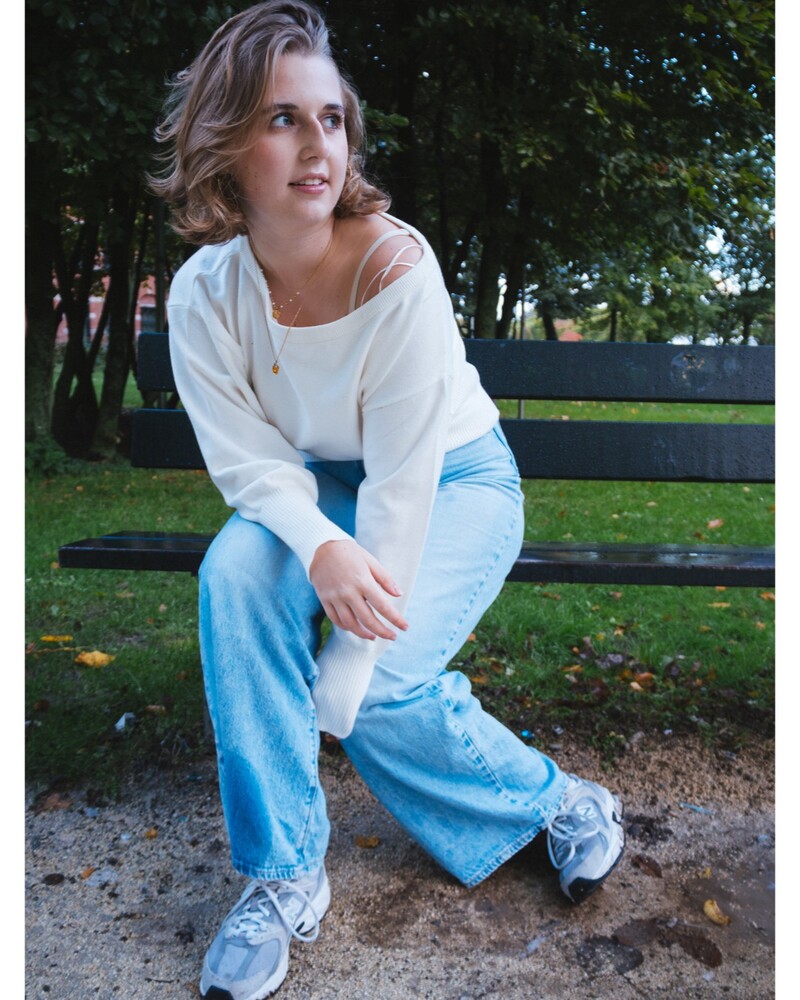 Young woman with shoulder-length wavy blonde hair sits on a park bench, looking to the side, wearing a cream off-shoulder sweater and light wash wide-leg jeans.