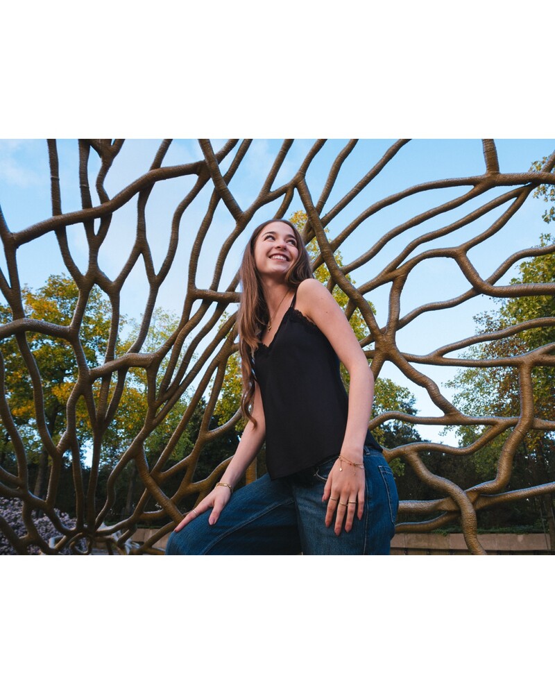 Smiling woman in a black sleeveless top and jeans sitting in a curved, branch-like metal lattice structure outdoors