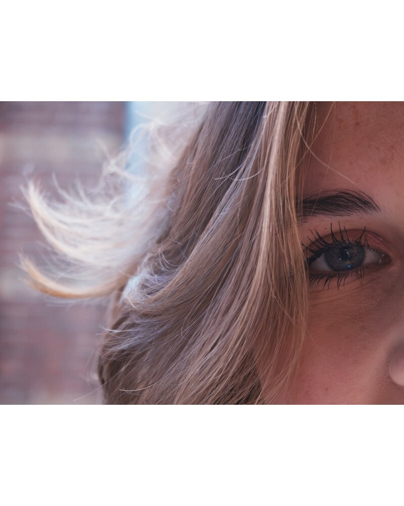 Close-up of a woman's face showing one blue-gray eye and blonde hair blowing across the frame, with a blurred brick background.
