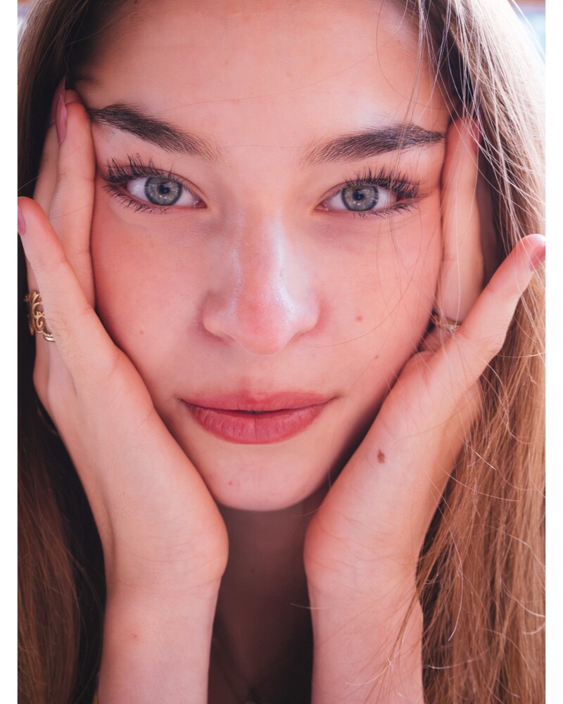 Close-up portrait of a young woman with light blue eyes and long lashes, her hands cupping both sides of her face.